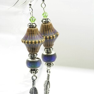 Pu&ograve; includere: Un paio di orecchini pendenti con un ciondolo in ceramica marrone e viola, una perla blu iridescente e un ciondolo a forma di piuma d'argento.