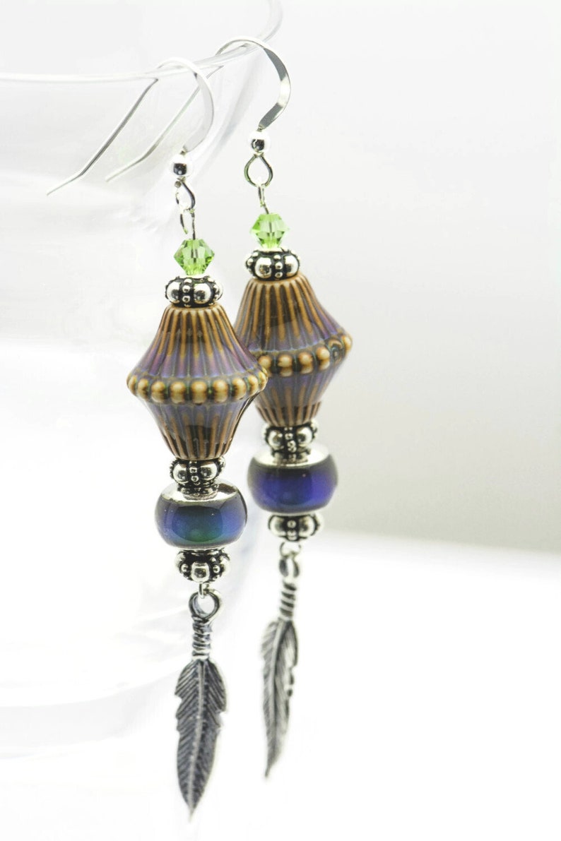 Pu&ograve; includere: Un paio di orecchini pendenti con un ciondolo in ceramica marrone e viola, una perla blu iridescente e un ciondolo a forma di piuma d'argento.