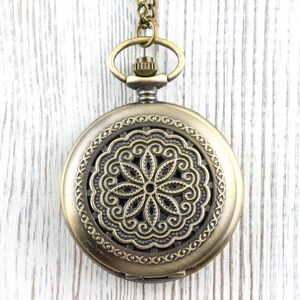 K&ouml;nnte beinhalten: Eine antike Taschenuhr aus Messing mit einem aufwendigen Blumendesign auf dem Zifferblatt. Die Uhr hat einen dekorativen Rand und h&auml;ngt an einer passenden Kette. Der Hintergrund ist eine wei&szlig;e Holzoberfl&auml;che.