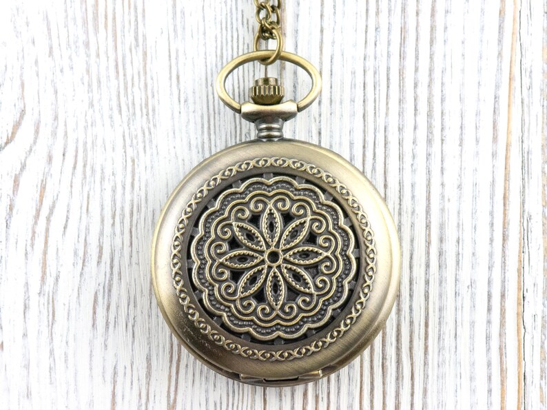K&ouml;nnte beinhalten: Eine antike Taschenuhr aus Messing mit einem aufwendigen Blumendesign auf dem Zifferblatt. Die Uhr hat einen dekorativen Rand und h&auml;ngt an einer passenden Kette. Der Hintergrund ist eine wei&szlig;e Holzoberfl&auml;che.