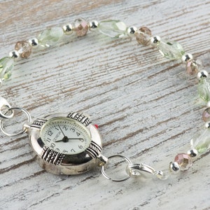 May include: A silver watch with a white face and black numbers. The watch is attached to a bracelet made of green and pink beads.