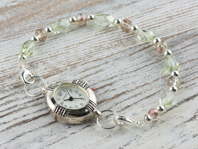 May include: A silver watch with a white face and black numbers. The watch is attached to a bracelet made of green and pink beads.