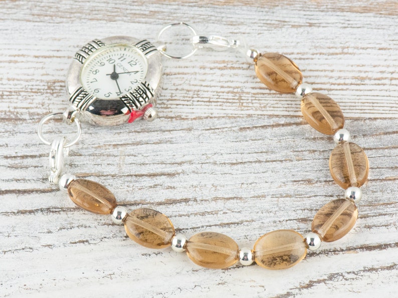 May include: A silver watch with a brown beaded bracelet. The watch face has black Roman numeral markings and a silver second hand.