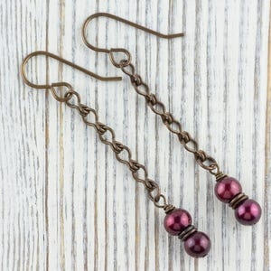 Puede incluir: Un par de pendientes largos colgantes con un dise&ntilde;o de cadena de bronce. Cada pendiente presenta una cadena que termina con dos cuentas redondas de color rojo oscuro. Los pendientes se exhiben sobre una superficie de madera blanca.