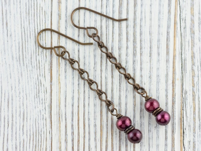 Puede incluir: Un par de pendientes largos colgantes con un dise&ntilde;o de cadena de bronce. Cada pendiente presenta una cadena que termina con dos cuentas redondas de color rojo oscuro. Los pendientes se exhiben sobre una superficie de madera blanca.