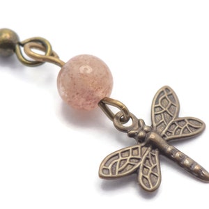 May include: A pair of earrings featuring a pink gemstone bead and a dragonfly charm. The dragonfly is made of bronze and has a detailed design.