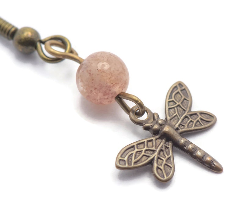 May include: A pair of earrings featuring a pink gemstone bead and a dragonfly charm. The dragonfly is made of bronze and has a detailed design.