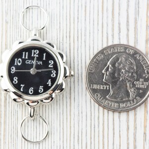 May include: A silver-toned watch with a black face and white numbers. The watch is about the size of a UK 20 pence coin.