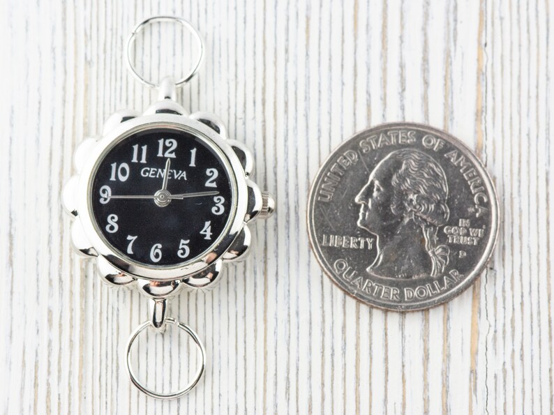 May include: A silver-toned watch with a black face and white numbers. The watch is about the size of a UK 20 pence coin.