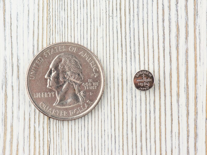 Pu&ograve; includere: Una moneta da un quarto di dollaro americano in argento e una batteria per orologio in argento si trovano su una superficie di legno bianco. La moneta presenta il profilo di George Washington e le parole "United States of America", "Liberty", "In God We Trust" e "Quarter Dollar". La batteria &egrave; etichettata con "SR626SW", "Maxell", "Hg 0" e "Japan".