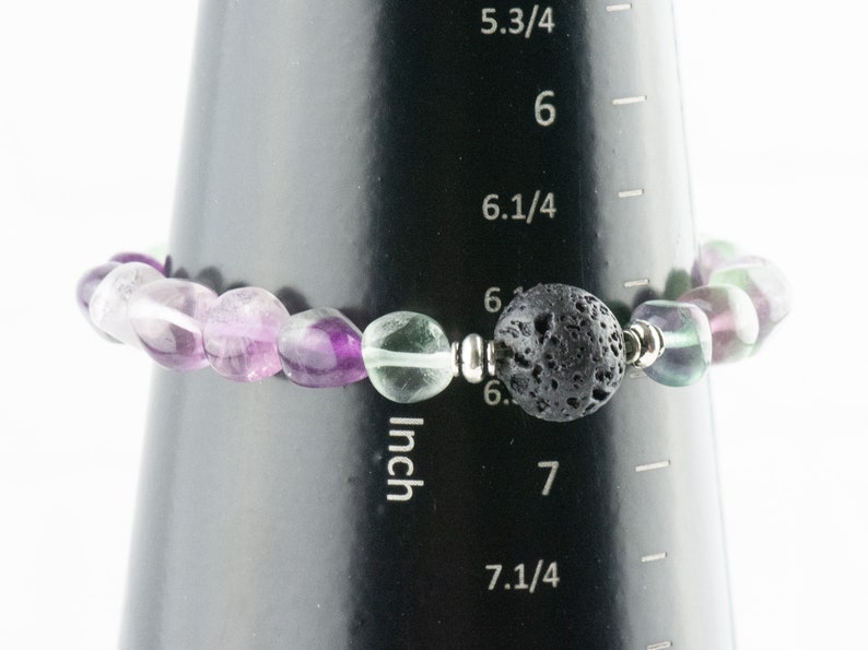 Puede incluir: Un brazalete de piedras preciosas de color morado, verde y blanco con una cuenta de lava negra y cuentas espaciadoras de plata. El brazalete mide 6,5 pulgadas de largo.