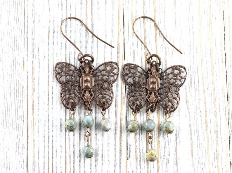 May include: A pair of bronze-colored butterfly earrings. The earrings feature intricate openwork wings and a central rectangular design. Each earring has three dangling, round, pale green and blue beads. The earrings hang from bronze-colored ear wires.