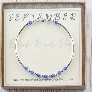 May include: A silver bangle bracelet with a blue crystal charm. The bracelet is in a box with the words "SEPTEMBER" and "Black Brook Shop" on it. The box also says "Nature-Inspired Jewellery and Watches".