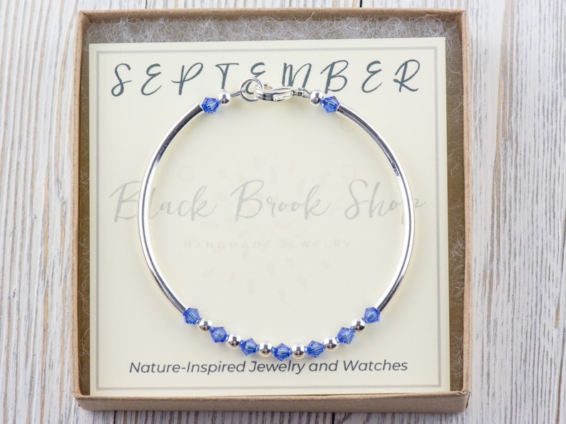 May include: A silver bangle bracelet with a blue crystal charm. The bracelet is in a box with the words "SEPTEMBER" and "Black Brook Shop" on it. The box also says "Nature-Inspired Jewellery and Watches".