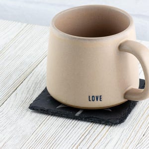 K&ouml;nnte beinhalten: Eine beige Keramiktasse mit einem schwarzen Henkel und dem Wort "LOVE" auf dem Boden. Die Tasse steht auf einem schwarzen Schieferuntersetzer.