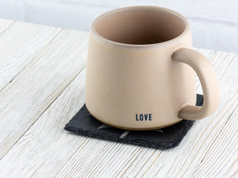 K&ouml;nnte beinhalten: Eine beige Keramiktasse mit einem schwarzen Henkel und dem Wort "LOVE" auf dem Boden. Die Tasse steht auf einem schwarzen Schieferuntersetzer.