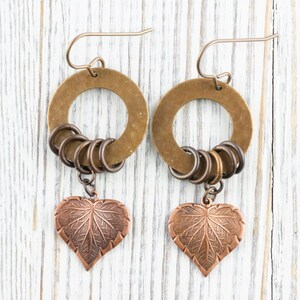 Op de afbeelding: Een paar handgemaakte oorbellen met een bronzen cirkelvormige ring, kleine bronzen ringen en een koperkleurige hartvormige bladhanger. De oorbellen worden gepresenteerd tegen een witte houten achtergrond.