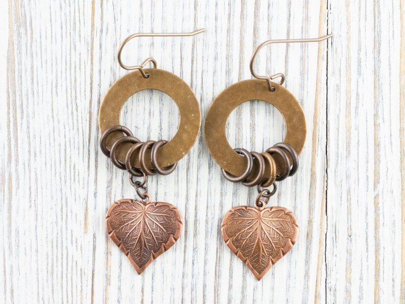 Op de afbeelding: Een paar handgemaakte oorbellen met een bronzen cirkelvormige ring, kleine bronzen ringen en een koperkleurige hartvormige bladhanger. De oorbellen worden gepresenteerd tegen een witte houten achtergrond.