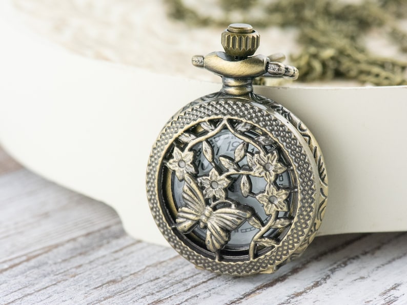 May include: A vintage-style pocket watch with a delicate butterfly design and floral accents. The watch face is visible through the openwork design, and the watch is attached to a chain.