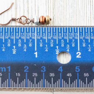 May include: Copper-colored earring with silver, copper, and gold-colored beads. The earring's hook is made of copper. The earring is approximately 1 inch (2.5 cm) long, as measured by the blue ruler in the image. The earring is on a light wood surface.