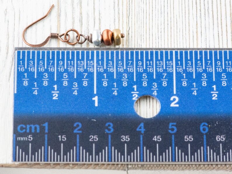 May include: Copper-colored earring with silver, copper, and gold-colored beads. The earring's hook is made of copper. The earring is approximately 1 inch (2.5 cm) long, as measured by the blue ruler in the image. The earring is on a light wood surface.