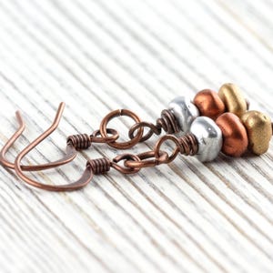 May include: Close-up of handmade earrings with copper-colored ear wires and chain links. Each earring features a cluster of metallic beads in silver, copper, and gold tones. The earrings rest on a white, wooden surface.