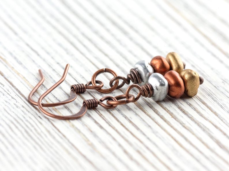May include: Close-up of handmade earrings with copper-colored ear wires and chain links. Each earring features a cluster of metallic beads in silver, copper, and gold tones. The earrings rest on a white, wooden surface.