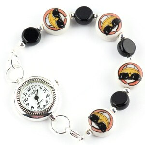 May include: A silver watch with a white face and black hands is attached to a bracelet made of black and white ceramic beads. The beads have a cartoon face with sunglasses on them.