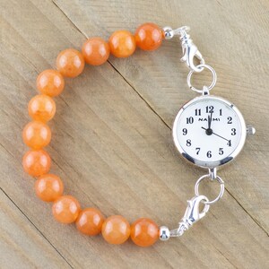 Op de afbeelding: Een zilveren zakhorloge met een witte wijzerplaat en zwarte cijfers is bevestigd aan een armband gemaakt van oranje kralen.