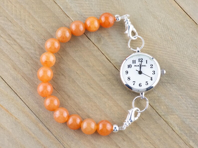 Op de afbeelding: Een zilveren zakhorloge met een witte wijzerplaat en zwarte cijfers is bevestigd aan een armband gemaakt van oranje kralen.