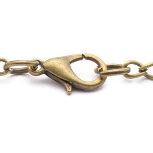 Puede incluir: Primer plano de una cadena de collar de color bronce con un cierre de mosquet&oacute;n. Los eslabones de la cadena son de forma ovalada y el cierre tiene un dise&ntilde;o curvo. El metal tiene un aspecto vintage y antiguo.