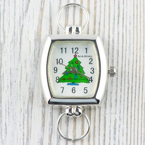 May include: Silver-toned Christmas-themed watch with a rectangular face. The watch face features a green Christmas tree illustration with red ornaments and a star. The watch has a ring at the top and bottom for attachment.