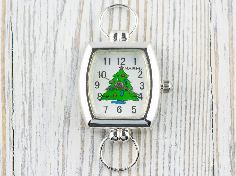 May include: Silver-toned Christmas-themed watch with a rectangular face. The watch face features a green Christmas tree illustration with red ornaments and a star. The watch has a ring at the top and bottom for attachment.