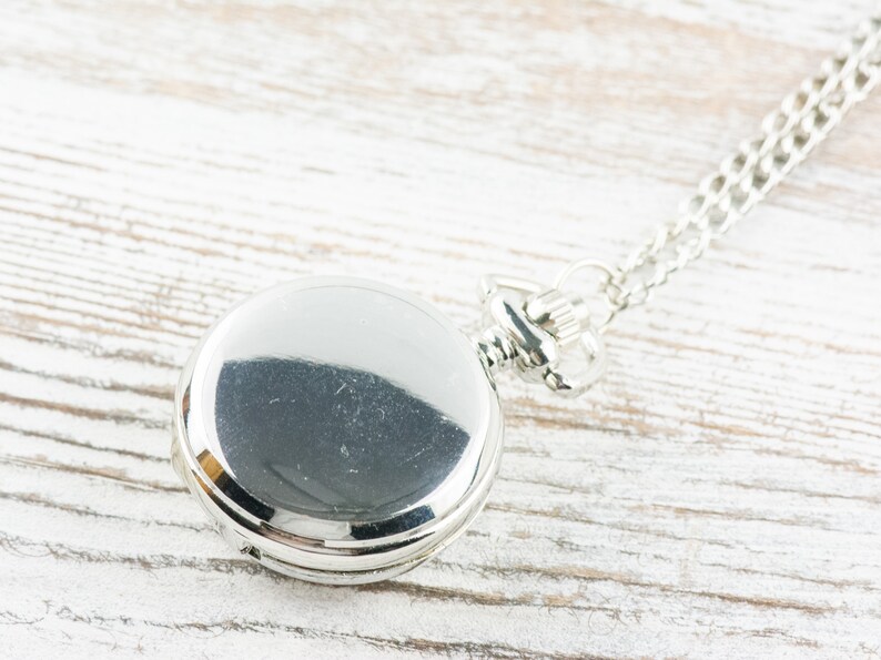 May include: A silver pocket watch with a chain on a white wooden surface.