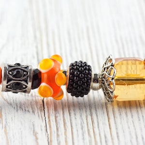 May include: Close-up of a bracelet with decorative beads. The beads include a black and silver bead, an orange and yellow glass bead, a black textured bead, and a yellow faceted bead with silver filigree.