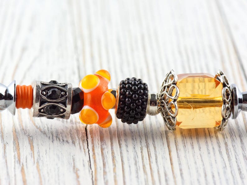 May include: Close-up of a bracelet with decorative beads. The beads include a black and silver bead, an orange and yellow glass bead, a black textured bead, and a yellow faceted bead with silver filigree.