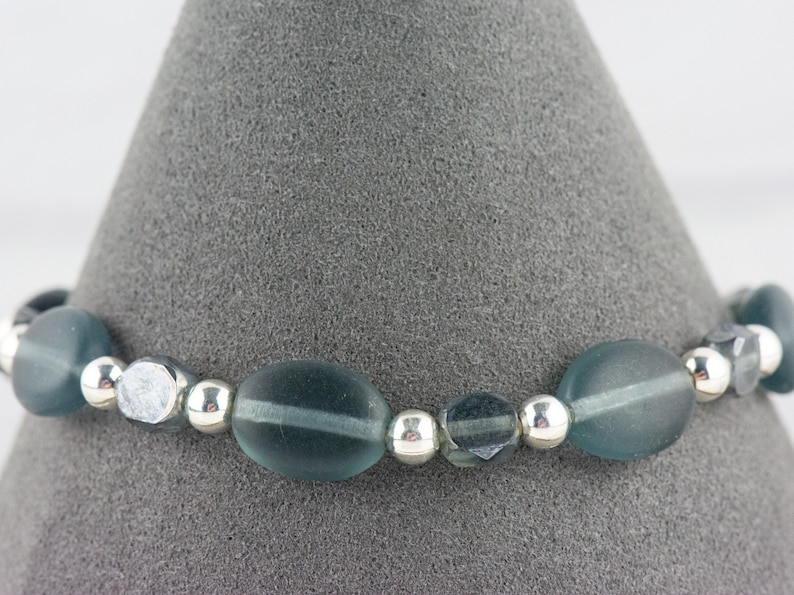 Pu&ograve; includere: Un braccialetto di perline con perline ovali e rotonde color teal intervallate da piccole perline d'argento lucido. Il braccialetto &egrave; esposto su uno sfondo grigio, mostrando il suo design e colore.