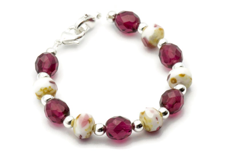 K&ouml;nnte beinhalten: Ein Armband mit abwechselnd burgunderfarbenen und wei&szlig;en Perlen. Die wei&szlig;en Perlen haben ein Blumenmuster. Das Armband hat einen silbernen Verschluss.