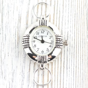 Pu&ograve; includere: Orologio da tasca Geneva argentato con quadrante bianco. L'orologio ha un quadrante rotondo con numeri e lancette nere. L'orologio &egrave; attaccato a un anello d'argento nella parte superiore e inferiore. L'orologio &egrave; su una superficie di legno bianco.
