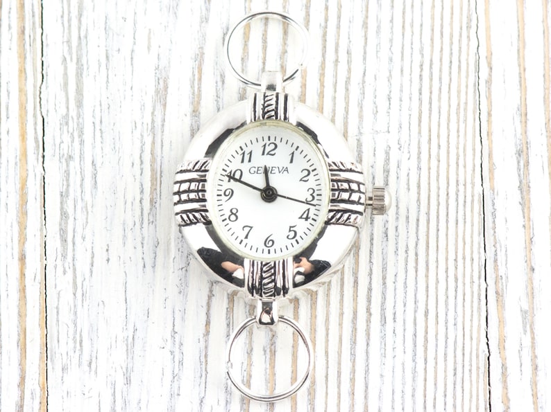 Pu&ograve; includere: Orologio da tasca Geneva argentato con quadrante bianco. L'orologio ha un quadrante rotondo con numeri e lancette nere. L'orologio &egrave; attaccato a un anello d'argento nella parte superiore e inferiore. L'orologio &egrave; su una superficie di legno bianco.