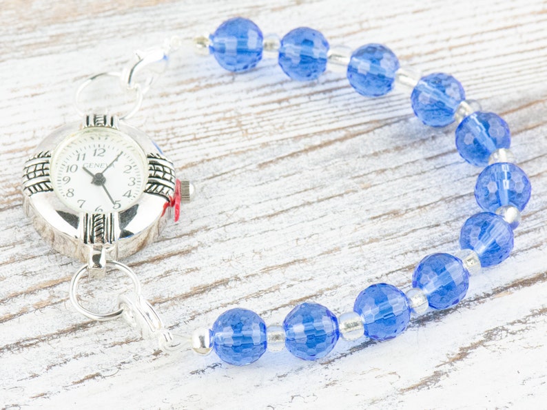 Pu&ograve; includere: Un orologio d'argento con quadrante bianco e numeri neri &egrave; attaccato a un braccialetto di perline blu. Il braccialetto &egrave; fatto di perline di vetro blu sfaccettate e distanziatori argentati.