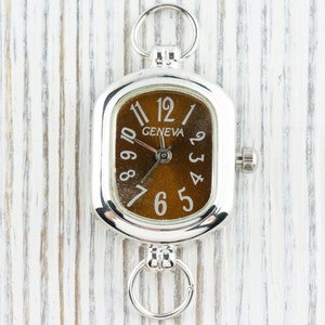 May include: A silver pocket watch with a rectangular face and a brown dial. The watch has the word "GENEVA" and white numerals. It has rings at the top and bottom for attachment.