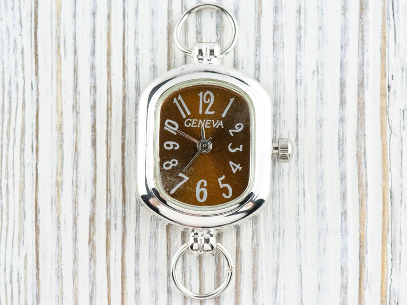 May include: A silver pocket watch with a rectangular face and a brown dial. The watch has the word "GENEVA" and white numerals. It has rings at the top and bottom for attachment.