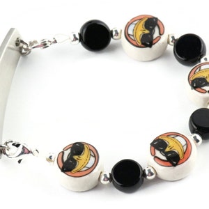 May include: A silver bracelet with a rectangular tag and black and white ceramic beads. The beads feature a cartoon character with sunglasses and a smile.