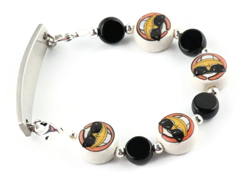 May include: A silver bracelet with a rectangular tag and black and white ceramic beads. The beads feature a cartoon character with sunglasses and a smile.