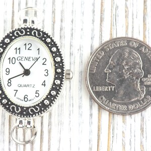 May include: Silver-toned Geneva Quartz watch with a white face and black numerals. The watch has an oval shape and a decorative bezel. A U.S. quarter is next to the watch for size comparison.