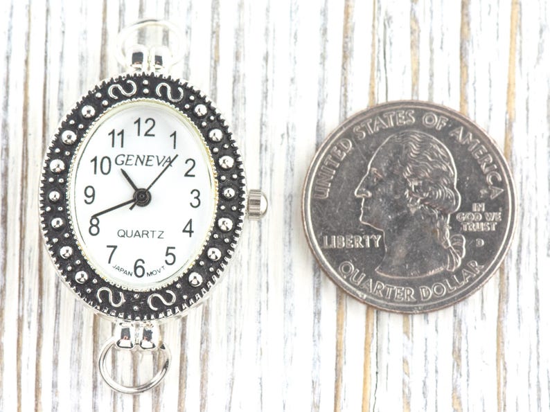 May include: Silver-toned Geneva Quartz watch with a white face and black numerals. The watch has an oval shape and a decorative bezel. A U.S. quarter is next to the watch for size comparison.