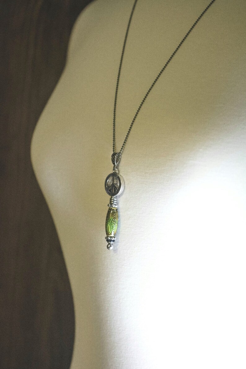 Puede incluir: Un collar de cadena de plata con un colgante de signo de paz y una cuenta verde y plateada.