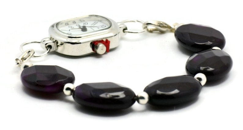 May include: A silver watch with a white face and black hands attached to a bracelet made of dark purple oval-shaped beads.