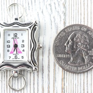 K&ouml;nnte beinhalten: Eine silberfarbene Geneva-Quarzuhr mit rosa Schleifendesign auf dem Zifferblatt, neben einer M&uuml;nze. Die Uhr hat ein rechteckiges Zifferblatt mit schwarzen Ziffern und Zeigern. Die Uhr ist etwa 2.5 cm lang.